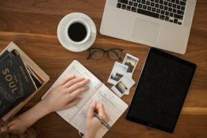 an aerial of a creative workspace featuring a ballpoint pen on an open notebook, books, a cup of coffee, a closed notebook, and a laptop on a desk.